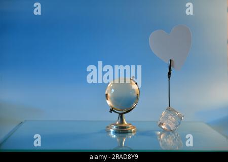 Glass globe and white paper heart on glass surface against blue background. Stock Photo