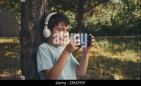 Excited happy schoolboy in wireless headphones playing online video game on smartphone, sitting under tree in the park Stock Photo