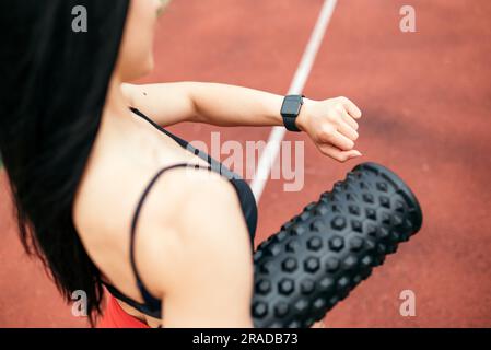 Sportswoman looking at smartwatch and holding massage roller in her other hand, outdoors. Fitness female setting up her smartwatch for her exercise. Stock Photo