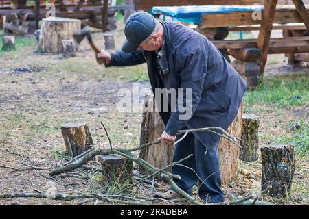 Elderly retired man chopping firewood with an ax, cooking grilled food on a fire Stock Photo