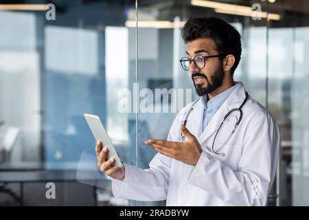 Successful professional Indian doctor working inside modern clinic, man consulting patients remotely, using tablet computer for video call, and long distance communication. Stock Photo