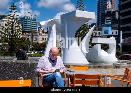 A man checks his mobile phone sat at a cafe by a fountain sculpture on Great Harbour Way in the Harbour Area, Wellington, Capital City, New Zealand Stock Photo