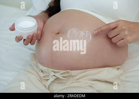 close up pregnant woman stroking apply heart shape cream on her belly on a bed Stock Photo