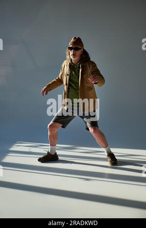 full length of expressive senior man in trendy hipster clothes and dark sunglasses standing and posing on grey background with lighting, stylish pose, Stock Photo