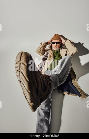 full length of cool and bearded senior man in dark sunglasses and beanie hat laying with outstretched leg on grey background with shadow, hipster styl Stock Photo