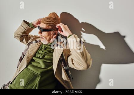 high angle view of senior bearded man in dark sunglasses and green hoodie adjusting beanie hat and looking away while laying on grey background with s Stock Photo