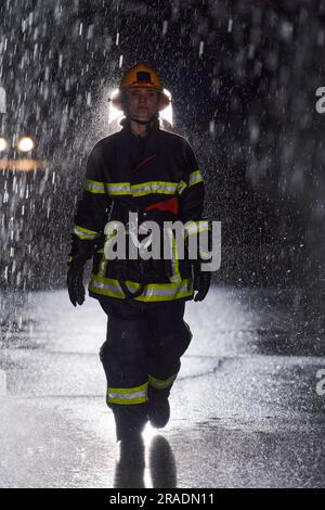 A determined female firefighter in a professional uniform striding through the dangerous, rainy night on a daring rescue mission, showcasing her Stock Photo