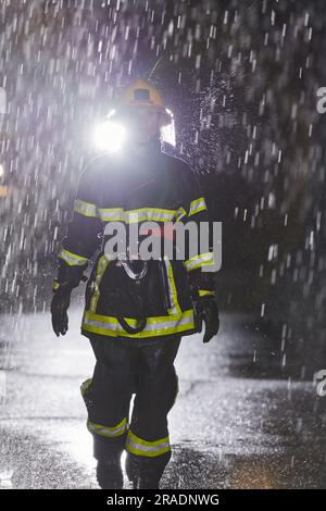 A determined female firefighter in a professional uniform striding through the dangerous, rainy night on a daring rescue mission, showcasing her Stock Photo