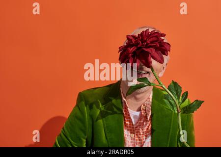 optimistic and trendy senior male model in green velour blazer hiding behind red peony while sitting on vibrant orange background, elderly man in gree Stock Photo