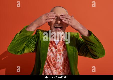 happy aging lifestyle, excited senior man smiling and covering eyes with hands while waiting for surprise on red orange background, green velour blaze Stock Photo