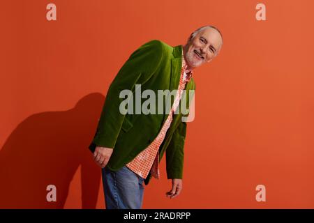 charismatic and carefree senior model posing on red orange background with shadow and smiling at camera, trendy casual clothes, green velour blazer, p Stock Photo