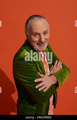 optimistic and charismatic bearded senior man standing with folded arms and smiling at camera on red orange background, fashion look, personal style, Stock Photo