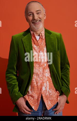 carefree, grey haired and fashionable senior man in green velour blazer and trendy shirt standing with thumbs in pockets and smiling at camera on red Stock Photo