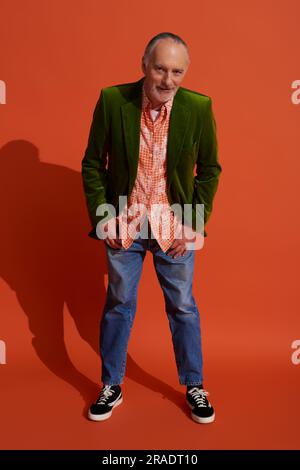 full length of positive elderly man in shirt and green velour blazer holding thumbs in pockets of blue jeans, leaning forward and looking at camera on Stock Photo