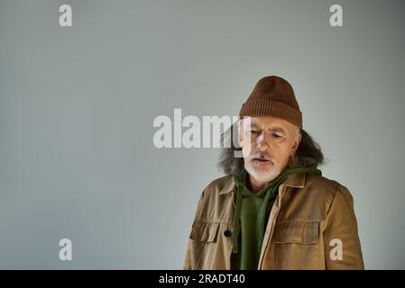 upset grey haired and bearded senior man in beanie hat and brown jacket standing on grey background, hipster style, expressive personality, aging popu Stock Photo