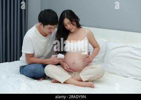 pregnant couple stroking apply heart shape cream on her belly on a bed Stock Photo