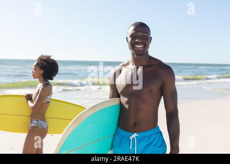 Portrait of happy african american man holding surfboard standing on sunny beach with female surfer Stock Photo