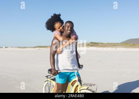 Happy african american couple embracing riding bike on sunny beach Stock Photo