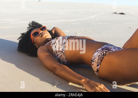 Happy african american woman in sunglasses and bikini sunbathing on sunny beach Stock Photo