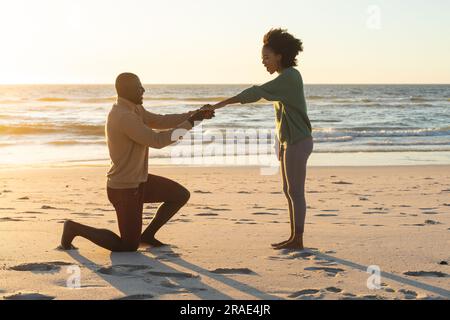 Happy african american man kneeling and proposing to girlfriend on beach at sundown Stock Photo