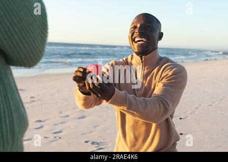 Happy african american man kneeling with ring and proposing to girlfriend on sunny beach Stock Photo