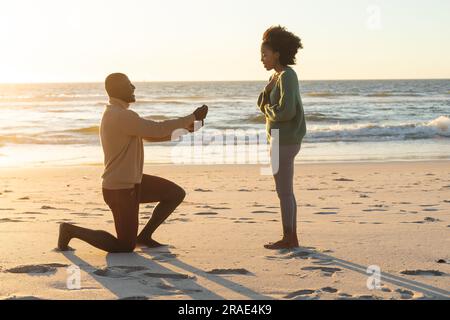 Happy african american man kneeling and proposing to girlfriend on beach at sundown Stock Photo