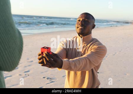 Happy african american man kneeling with ring and proposing to girlfriend on sunny beach Stock Photo