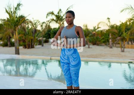 Happy african american woman practicing yoga meditation standing by pool on beach at sundown Stock Photo