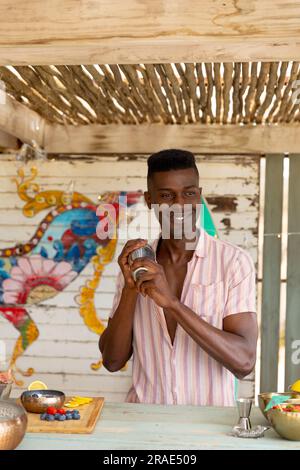 Happy african american male bartender preparing cocktail with shaker behind the counter at beach bar Stock Photo