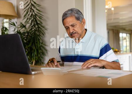 Senior biracial man doing paperwork and using laptop in dining room Stock Photo