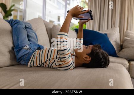 Happy biracial boy lying on couch with feet up using tablet in living room Stock Photo