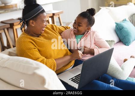 Happy african american mother and daughter sitting on sofa, embracing and using laptop at home Stock Photo