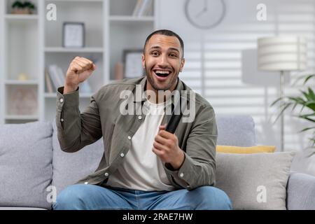 Portrait of a young African-American man sitting at home in front of the screen, holding the remote control in his hands and enthusiastically watching the game, competition. Happy shouts and cheers. Stock Photo