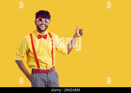 Young black haired Indian man in funny glasses for party shows thumb up stands on yellow background Stock Photo