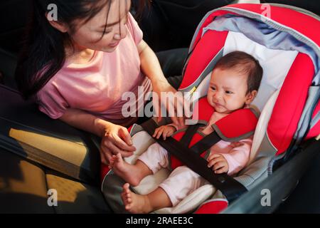 mother consoling her newborn baby crying while putting and fasten seat belts on car seat Stock Photo
