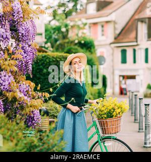 Retro styled portrait of beautiful young woman, wearing vintage clothes, holding mint color bicycle with yellow flowers placed in basket Stock Photo