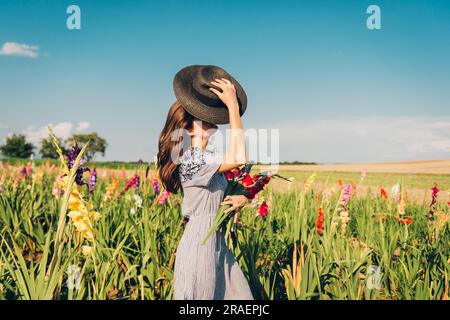 Gorgeous young woman picking flowers in a field, wearing summer dress, black straw hat Stock Photo
