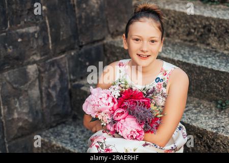 Summer portrait of adorable little girl wearing beautiful occasional dress, holding big peonies bouquet, posing outside Stock Photo