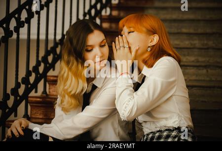 A friendship between two schoolgirls in uniform sitting on a staircase. A ginger Asian girl is whispering a secret into the ear of blond girlfriend. T Stock Photo