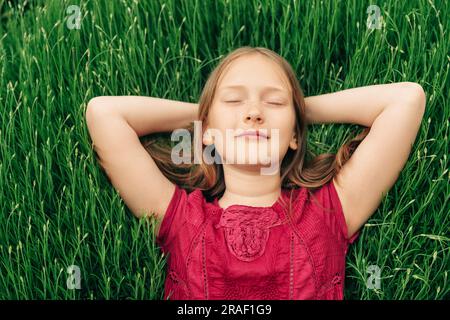 Cute little girl lying on bright green grass, holding arms behind head, eyes closed Stock Photo