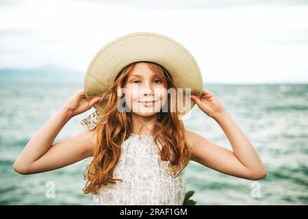 Outdoor portrait of pretty young kid girl with red hair, wearing big hat, family vacation with children by the sea Stock Photo