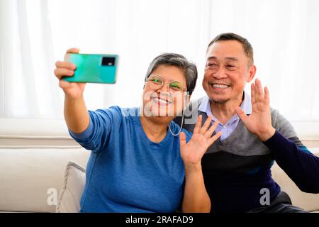 Portrait of healthy senior man and woman taking selfie photos on sofa at home, Happy senior married couple spending time in living room, Grandparents Stock Photo