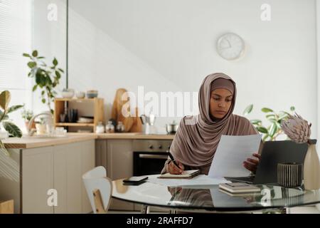 Young female employee reading financial document or contract and making notes while working over business project in home office Stock Photo