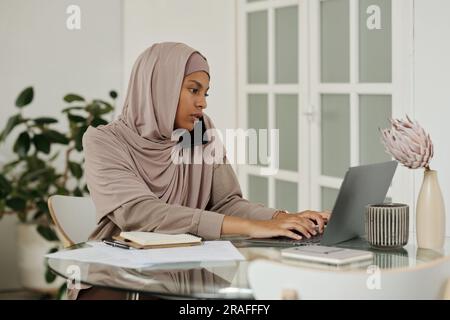 Young busy Muslim female employee with mobile phone by ear typing on laptop keyboard while organizing work by table in home environment Stock Photo