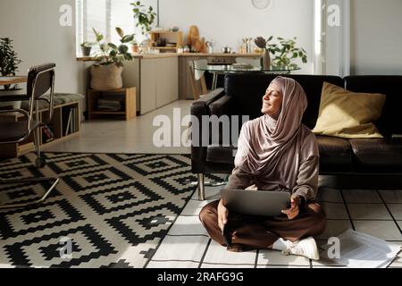 Young smiling woman in hijab using laptop and enjoying sunlight while sitting on the floor of living room and looking through window Stock Photo