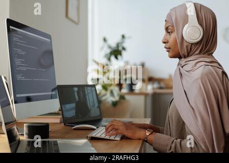 Side view of young female programmer in headphones and hijab decoding online data while communicating in video chat with colleague Stock Photo