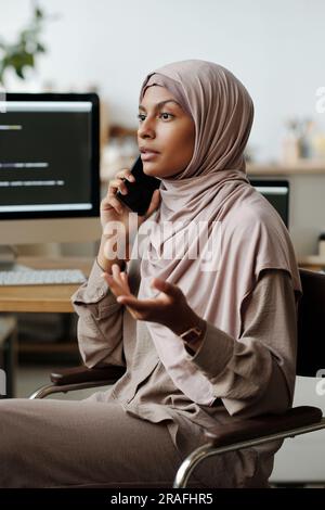 Young confident female IT engineer or programmer in hijab talking on smartphone while sitting by workplace in front of camera at home Stock Photo