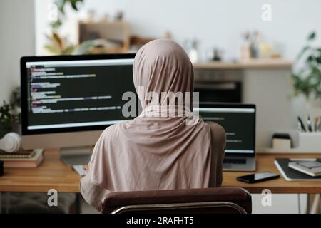 Rear view of Muslim female programmer in hijab sitting in front of computer screen with coded data and using new decoding software Stock Photo