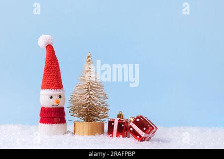 Knitted snowman in a red Christmas hat and sweater with two red gifts under the toy Christmas tree . Merry Christmas and New Year 2024 greeting card. Stock Photo