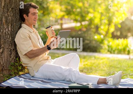 Relaxed guy sitting under tree at park, using digital tablet Stock Photo